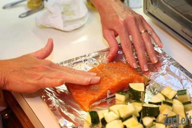 rubbing oil over a serving of salmon next to diced zucchini on a sheet pan.