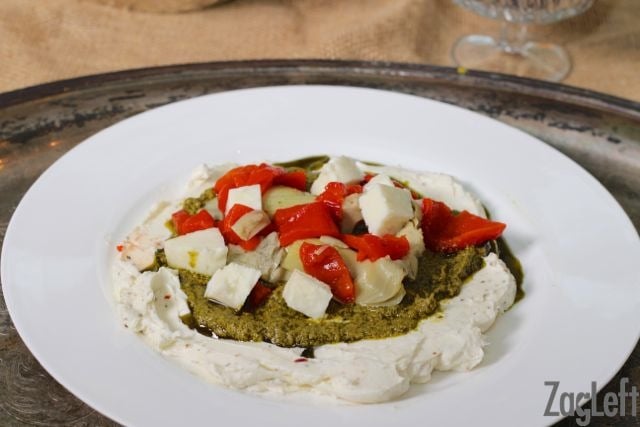 Layered Italian Cream Cheese Dip topped with pesto, artichoke hearts, red peppers and mozzarella on a plate