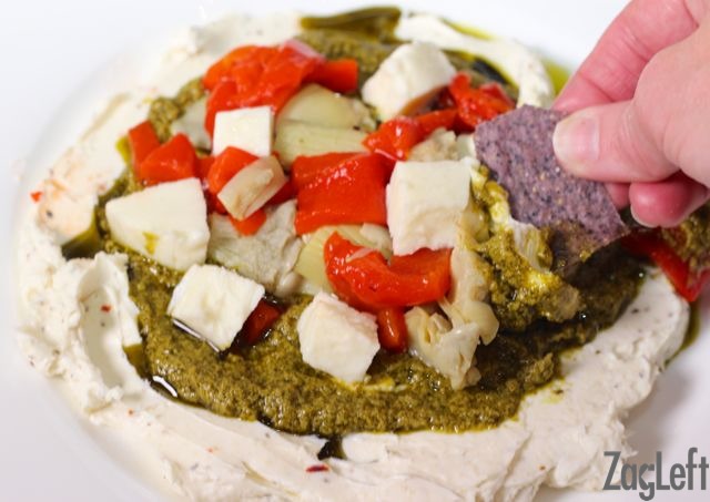 A blue corn tortilla chip dipped in the Layered Italian Cream Cheese Dip that's topped with pesto, artichoke hearts, red peppers, and mozzarella