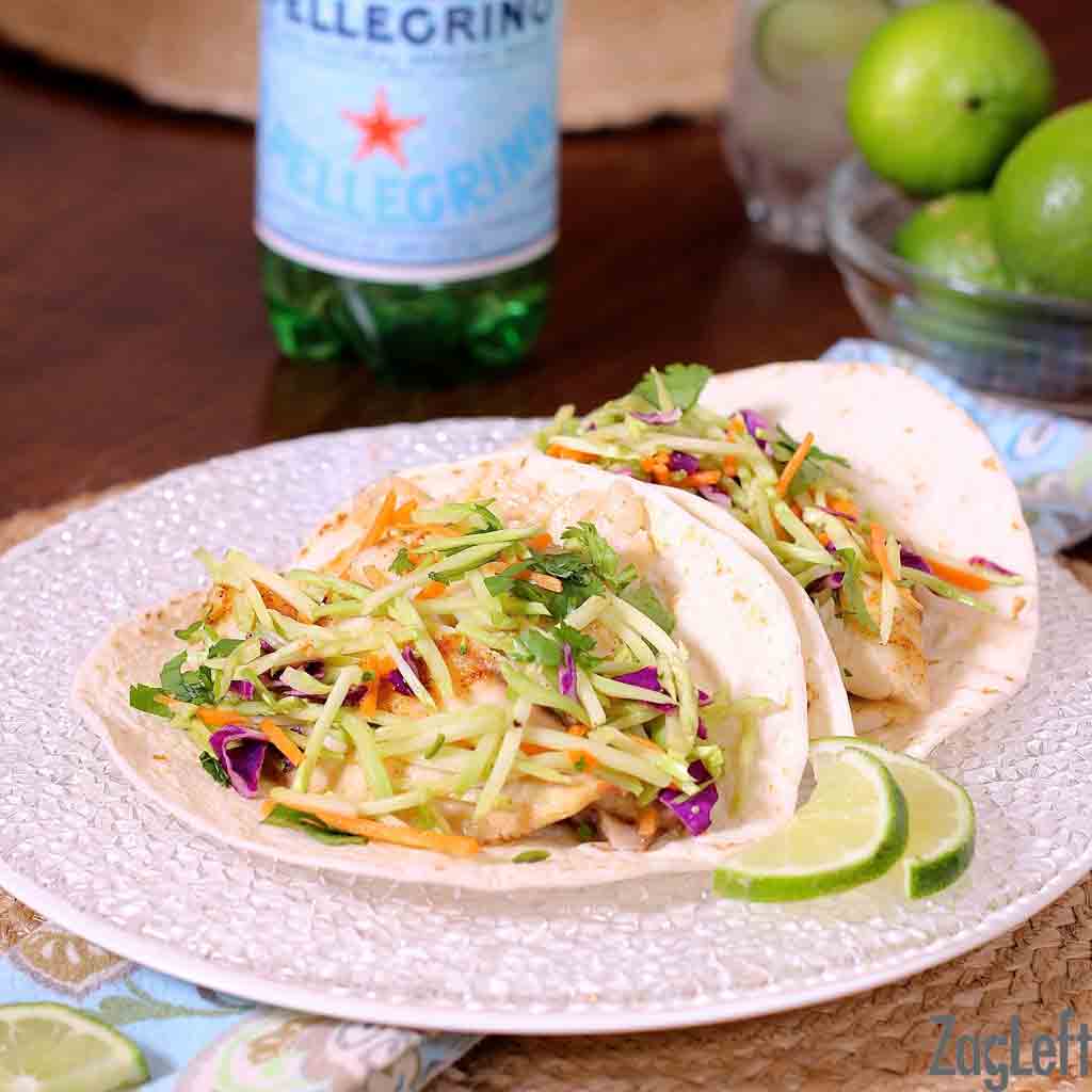 Two fish tacos topped with a broccoli slaw on a plate surrounded by fresh lime slices.