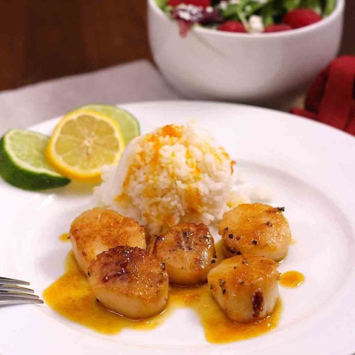 Sweet citrus sauce served over scallops and white rice on a plate garnished with a lemon and two lime wheels with a small salad in the background