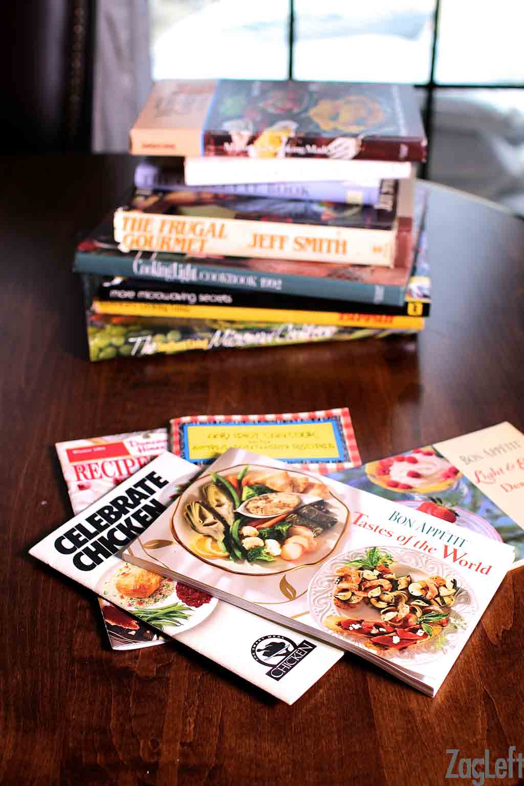 Five vintage cookbooks on a wooden table with a stack of cookbooks behind them