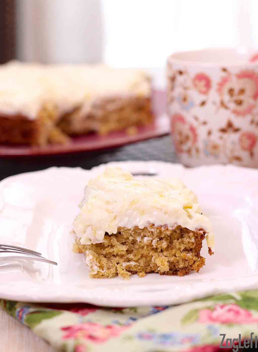 Closeup of a slice of pineapple oat cake with cream cheese frosting on a plate with a floral napkin and a coffee mug in the background.