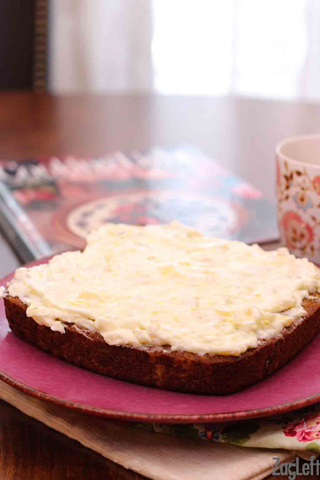 A closeup of a square cake frosted on a wooden table next to a cookbook and a coffee mug