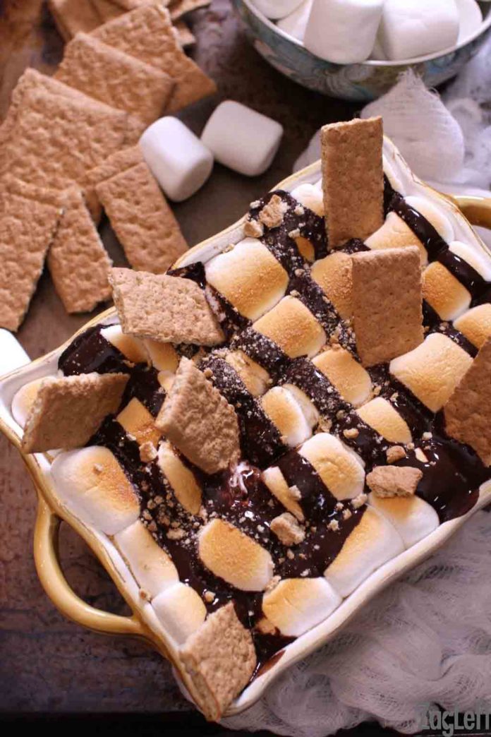 Overhead view of fudge brownies topped with marshmallows, graham crackers, and hot fudge in a baking dish with a bowl of marshmallows and graham crackers laid out behind the dish