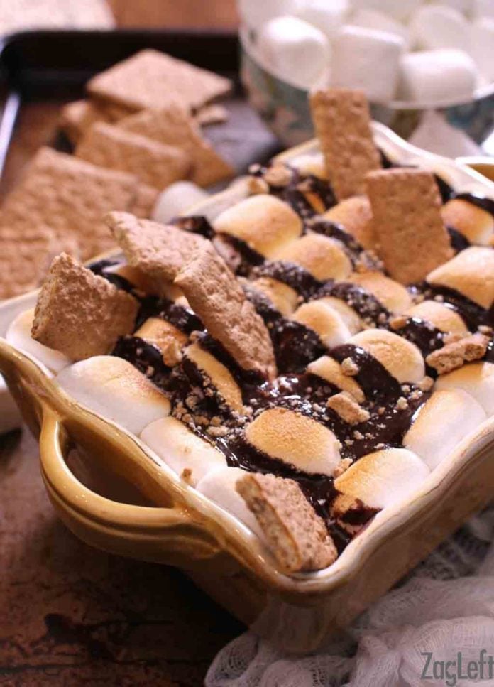 Fudge brownies topped with marshmallows, hot fudge, and graham crackers in a baking dish with a bowl of large marshmallows in the background 