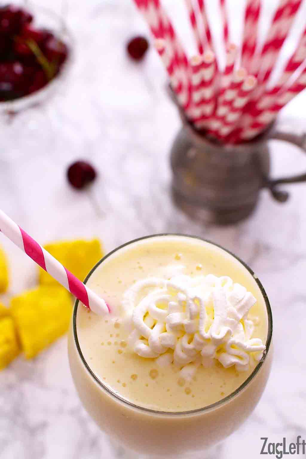 An overhead view of a smoothie topped with whipped cream next to a metal jar of red and white straws and a a few sliced pineapple pieces 