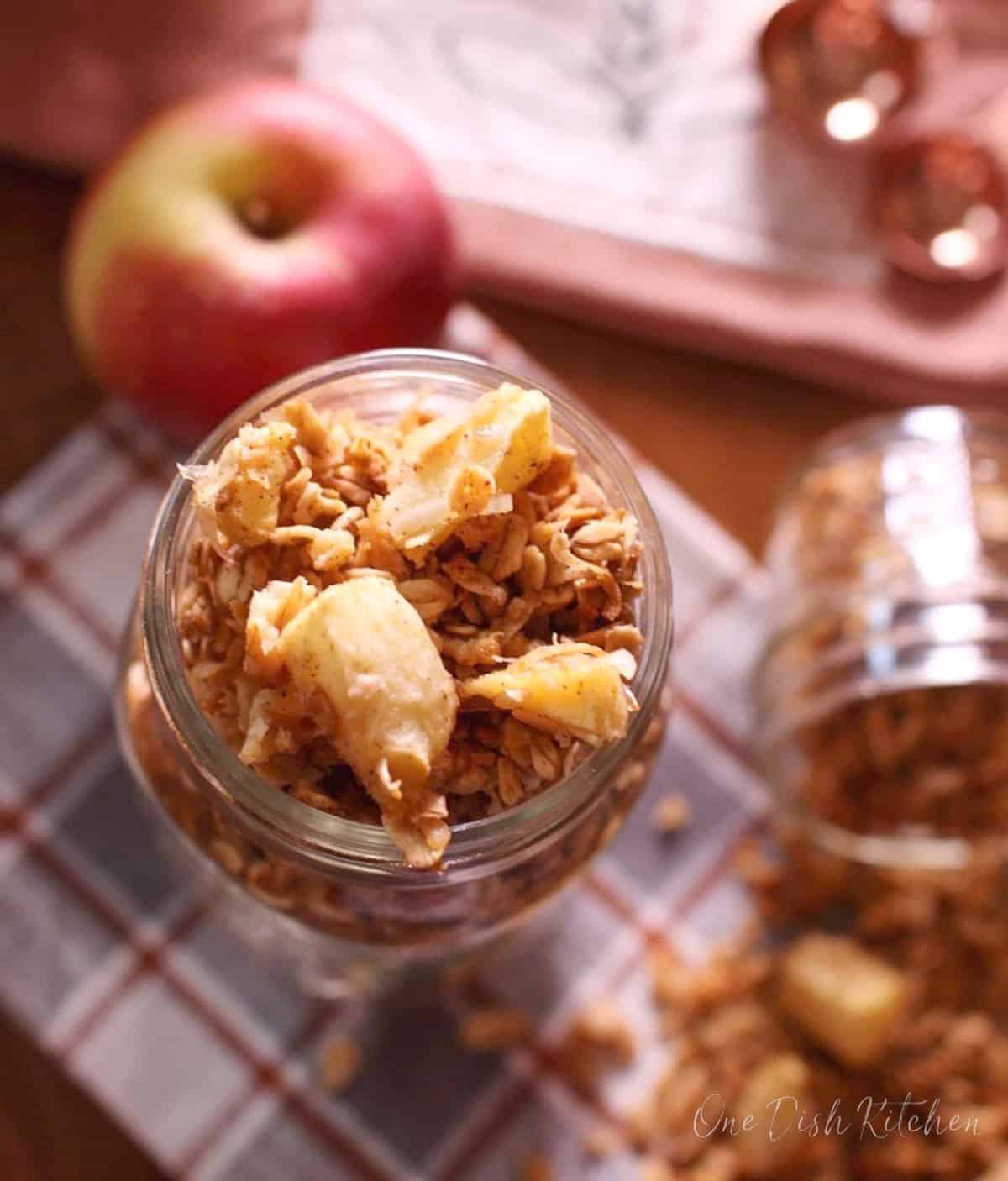 an overflowing jar of apple granola next to a spilled jar of granola on a table.