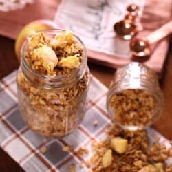 a jar of fresh apple granola on top of a plaid napkin and next to apples and measuring spoons