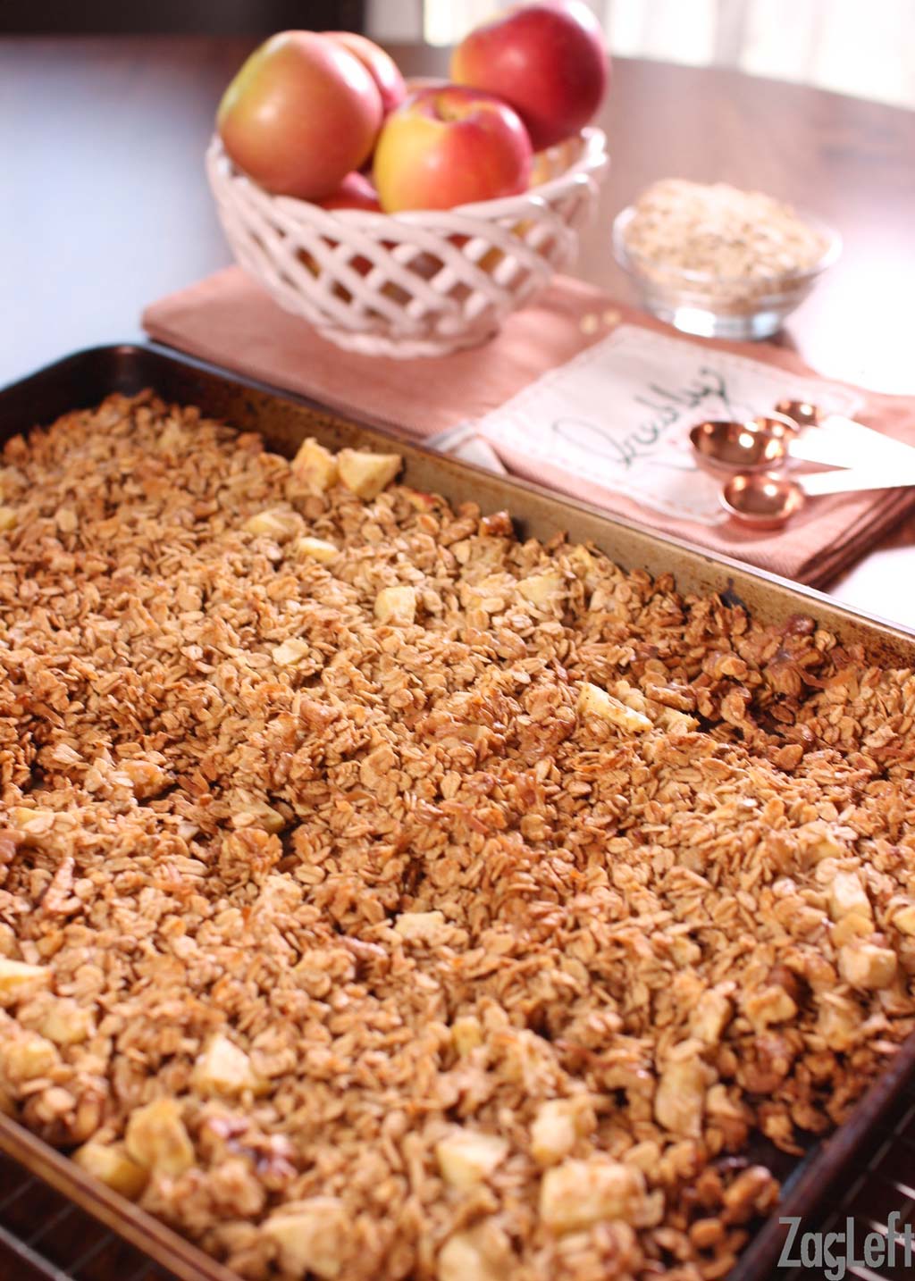 Apple granola on a baking sheet with a bowl of fresh apples, a small bowl of oats, and measuring spoons in the background.