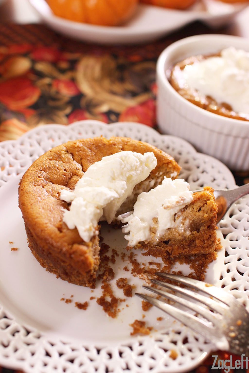 A forkful of pumpkin pie topped with whipped cream on a plate and another pumpkin pie in a ramekin topped with whipped cream in the background