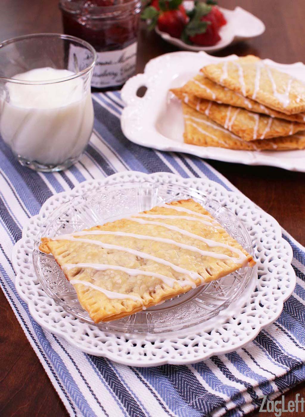 Homemade toaster pastries with icing stripes on a plate next to a glass of milk and a jar of jam.