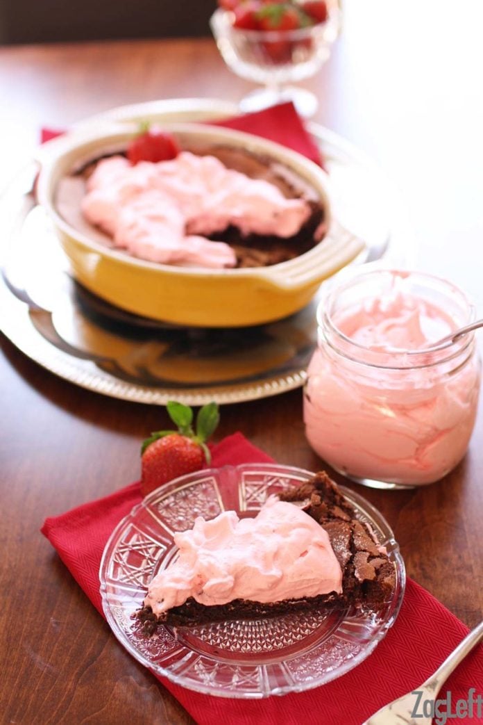Slice of flourless chocolate cake topped with strawberry whipped cream on a small plate with a jar of strawberry whipped cream and the remainder of chocolate cake topped with strawberry whipped cream in a baking dish in the background 