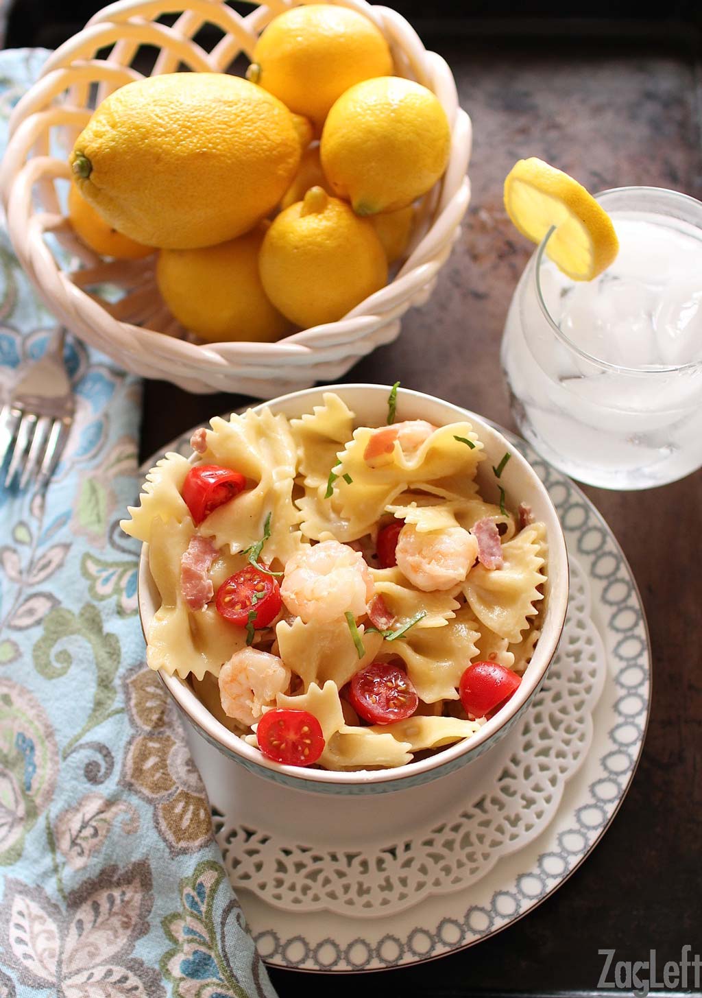 An overhead view of shrimp and prosciutto pasta in a small bowl plated on a metal tray with a small bowl of lemons and a glass of ice water.