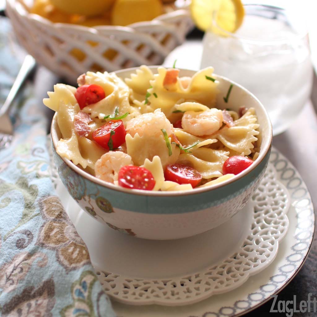 Shrimp and prosciutto pasta in a small bowl plated on a metal tray with a small bowl of lemons and a glass of ice water.