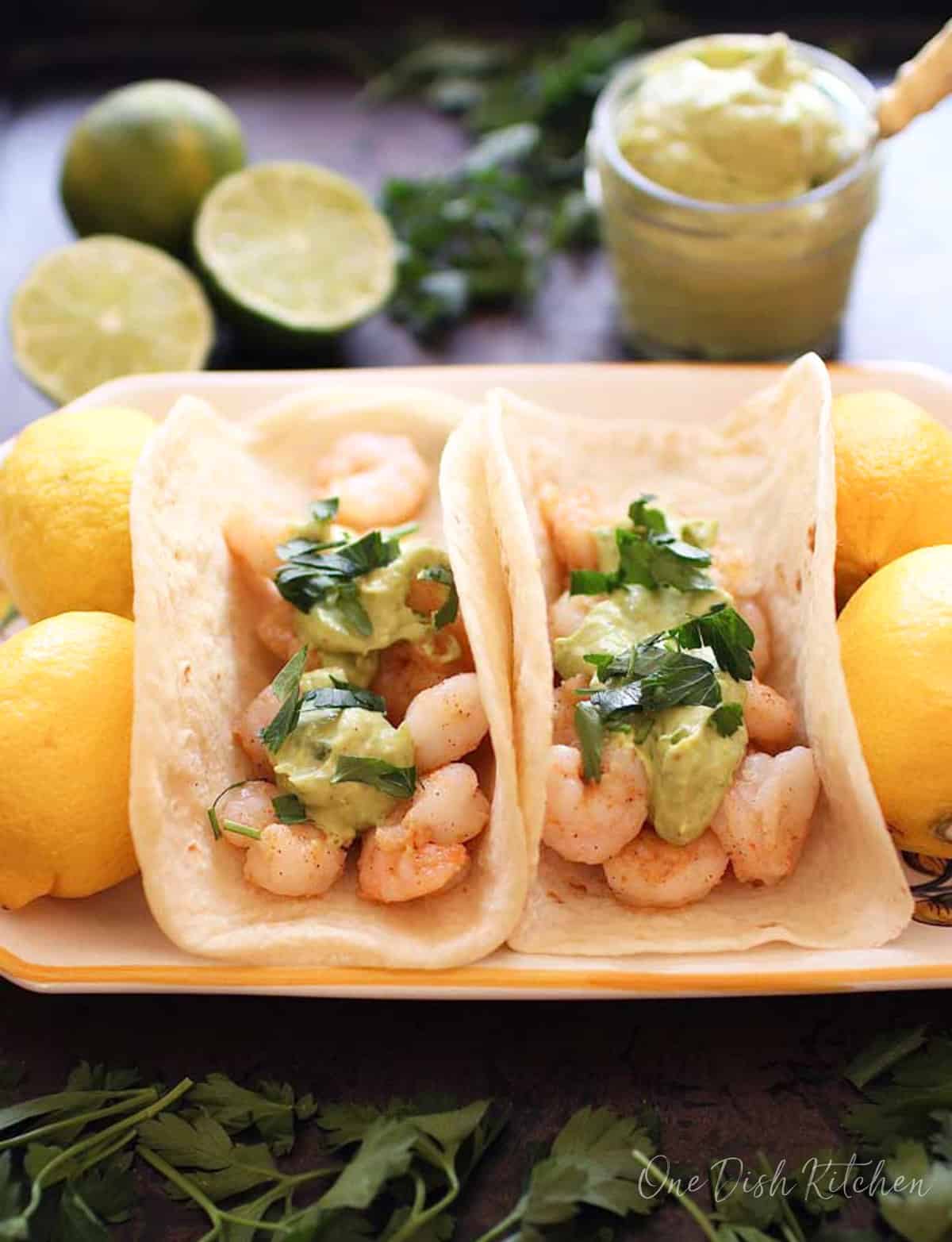 two shrimp tacos topped with avocado sauce on a plate next to four whole lemons