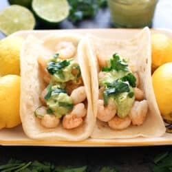two shrimp tacos with taco sauce on a plate next to lemons and limes