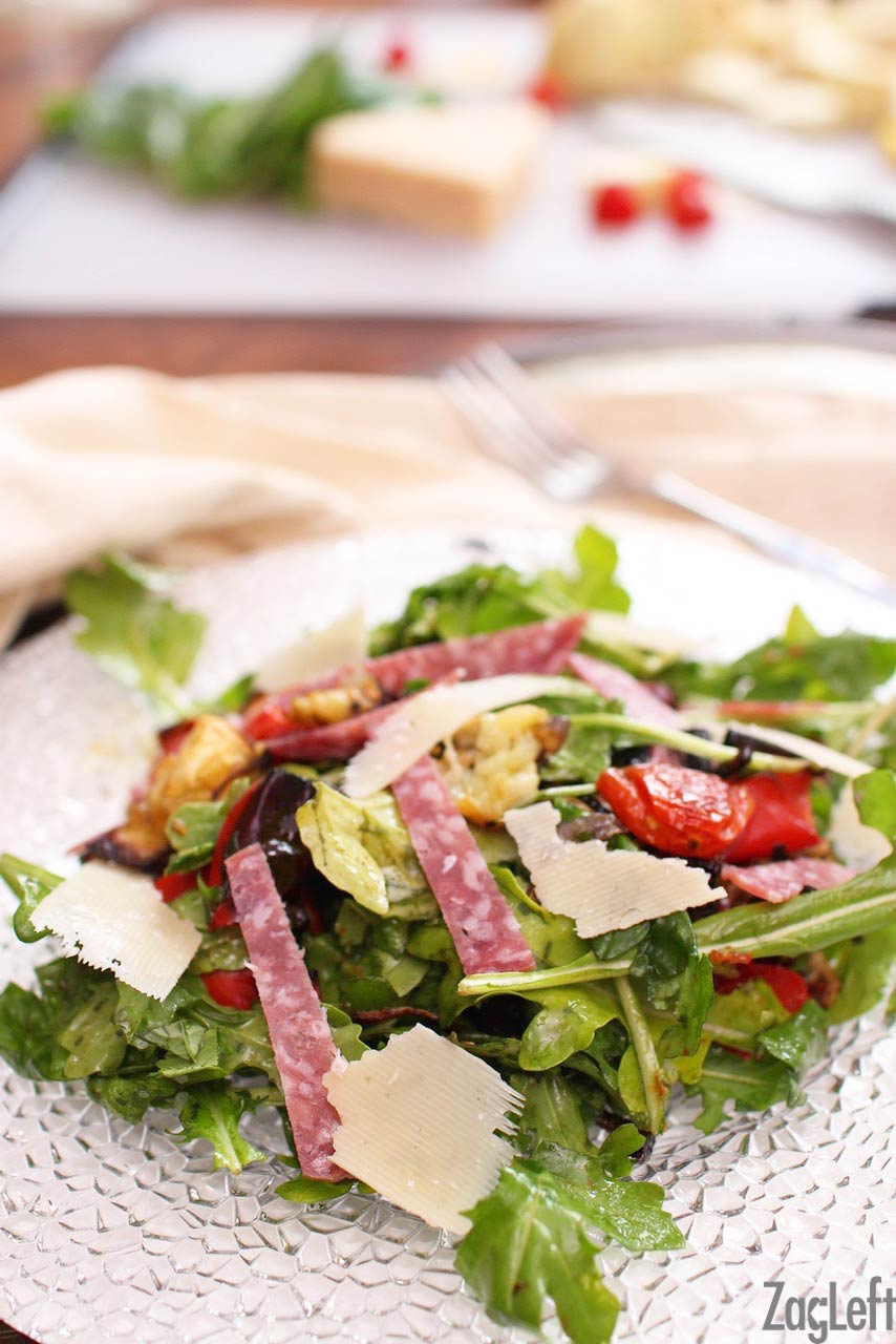 Antipasto salad made with spinach and topped with sliced salami strips, cherry tomatoes in halves, and sliced parmesan cheese on a plate.