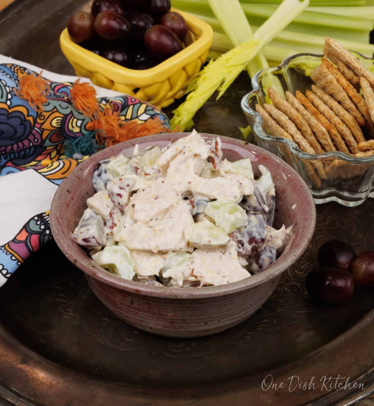 a pink bowl filled with chicken salad on a silver tray next to a bowl of crackers and a bowl of red grapes