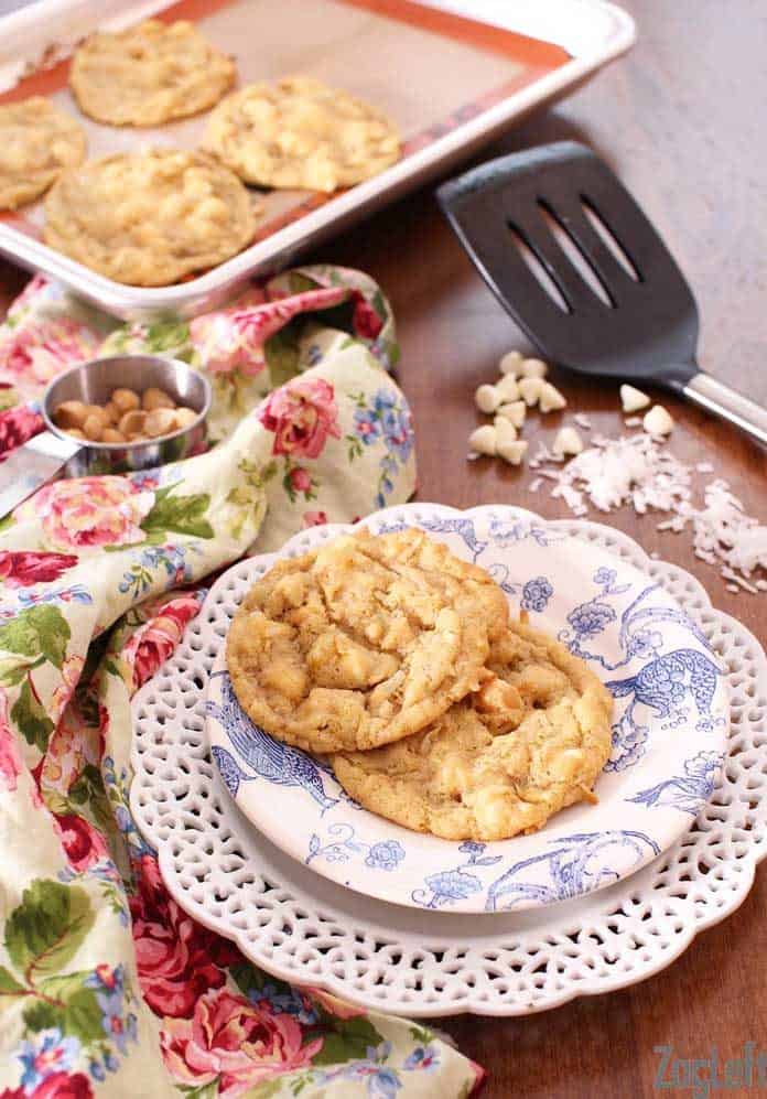Two Tropical White Chocolate Chip Cookies on a small plate next to a floral napkin and a tray of four cookies and spatula behind it
