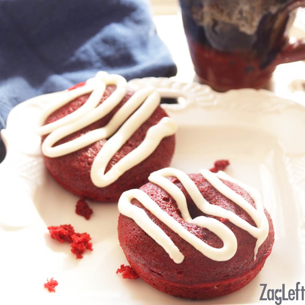 Two red velvet donuts topped with drizzled white frosting on a plate next to a cup of coffee and a blue napkin.