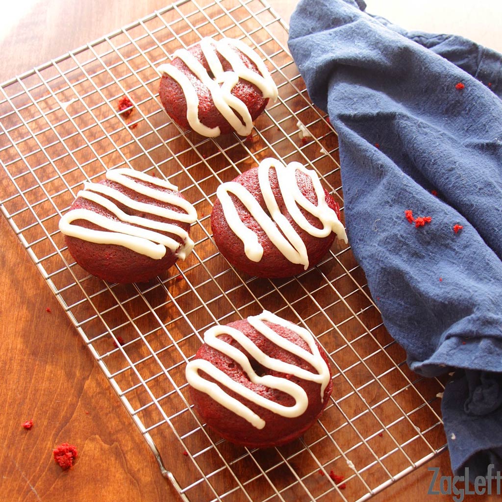 Overhead view of four donuts topped with drizzled white frosting on a cooling rack next to a blue cloth napkin.