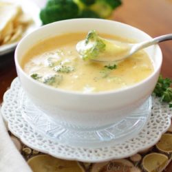a white bowl filled with broccoli cheese soup with crackers on a plate nearby