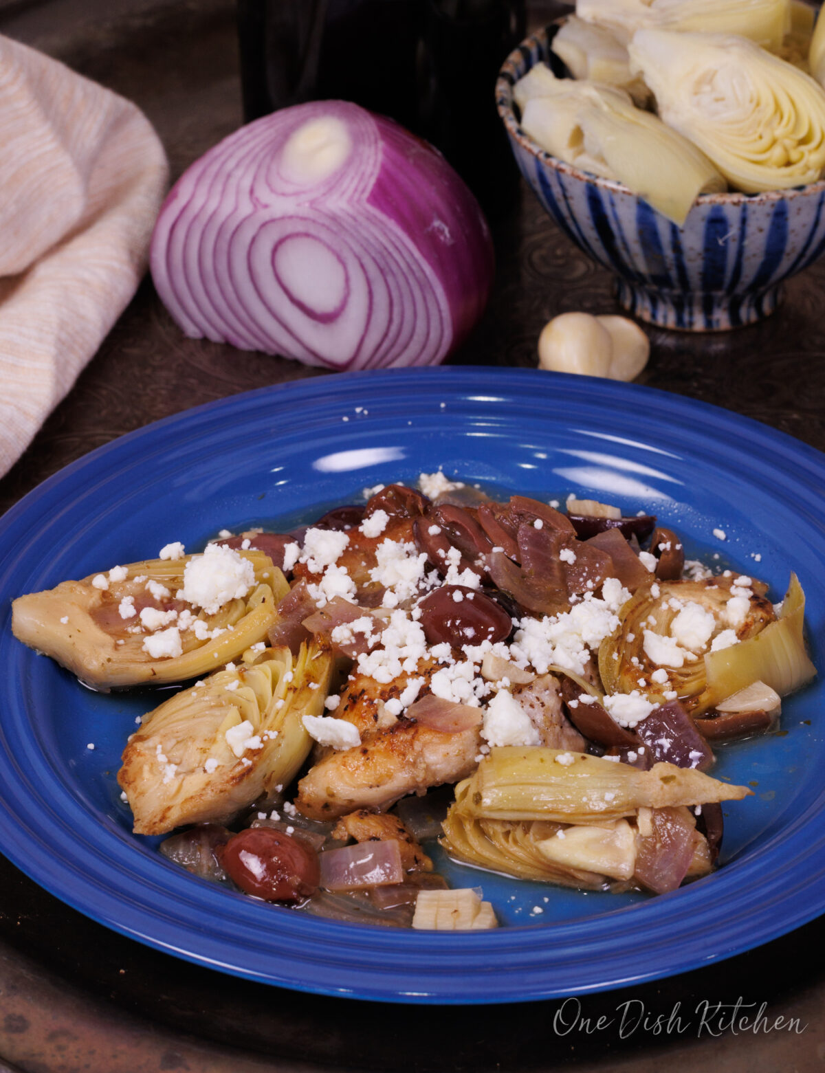 a chicken thigh on a blue plate surrounded by artichokes, red onions, and olives.
