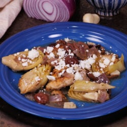 a chicken thigh on a blue plate surrounded by artichokes, red onions, and olives.