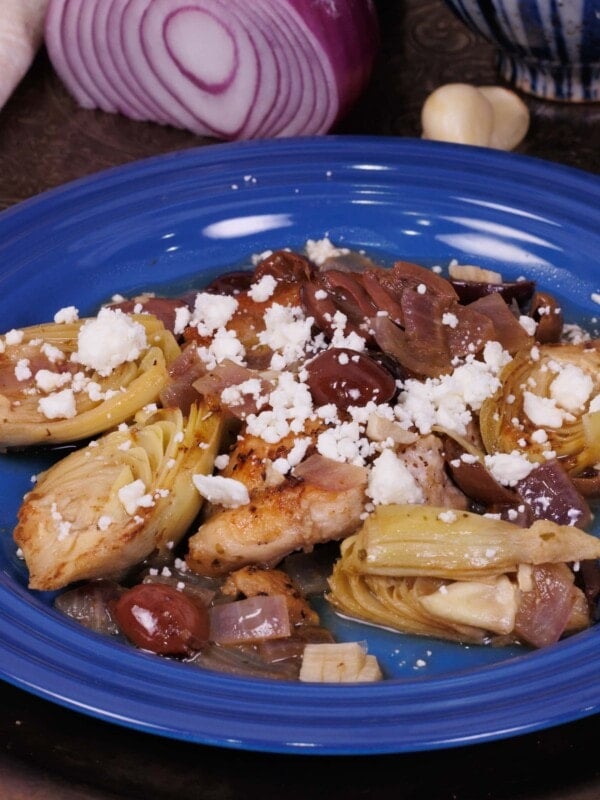 a chicken thigh on a blue plate surrounded by artichokes, red onions, and olives.