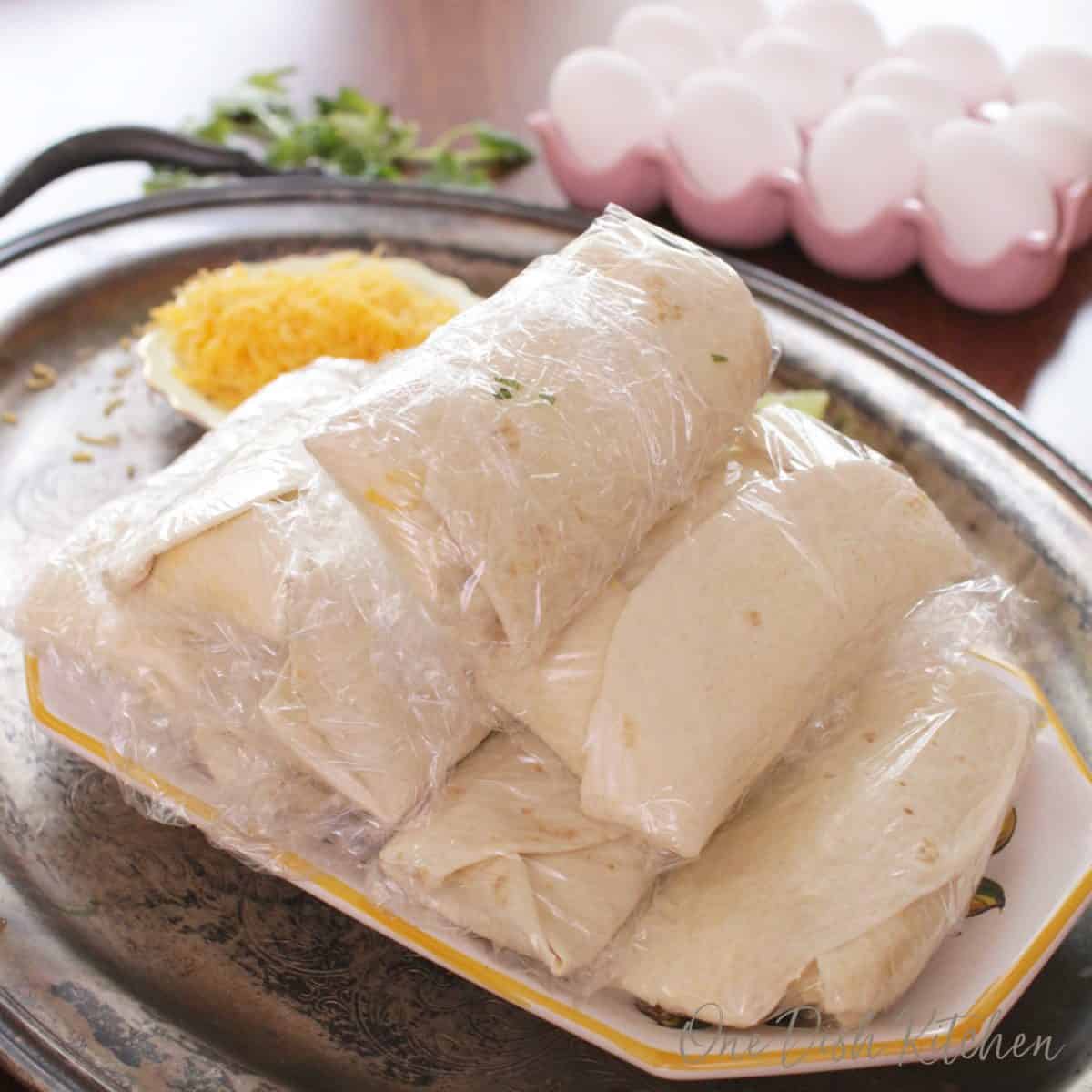 burritos individually wrapped in plastic wrap on a plate next to a pink tray filled with eggs.