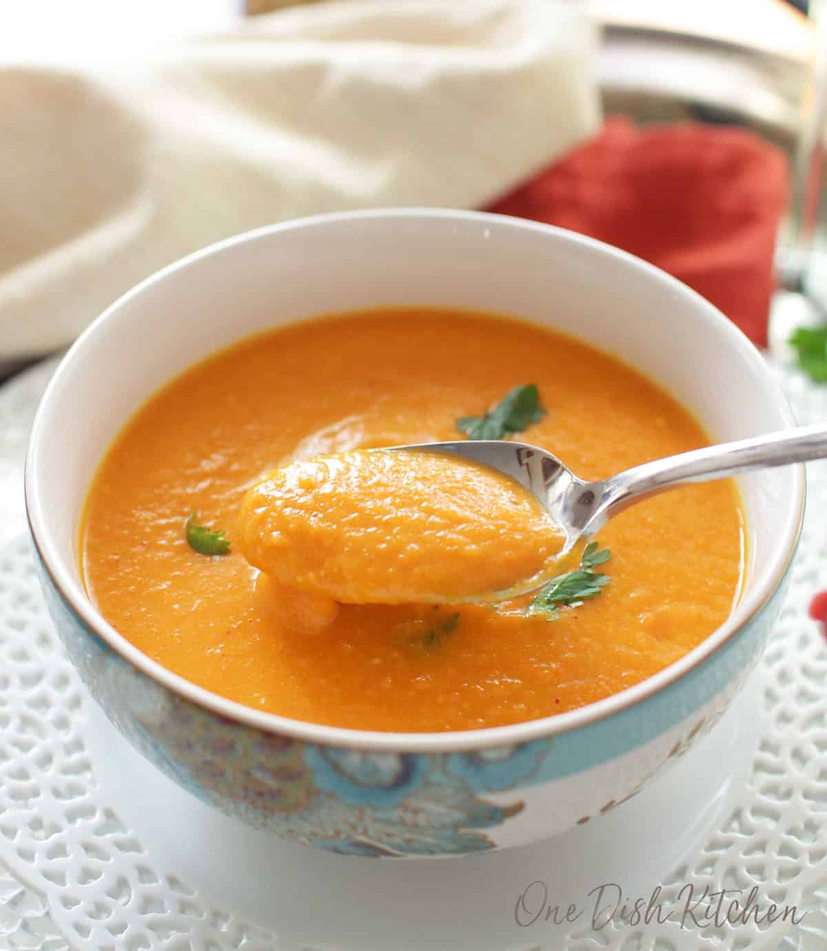 A bowl of carrot soup garnished with cilantro on a glass tray next to a glass of white wine and a bunch of cilantro.