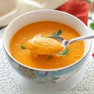 A bowl of carrot soup garnished with cilantro on a glass tray next to a glass of white wine and a bunch of cilantro.