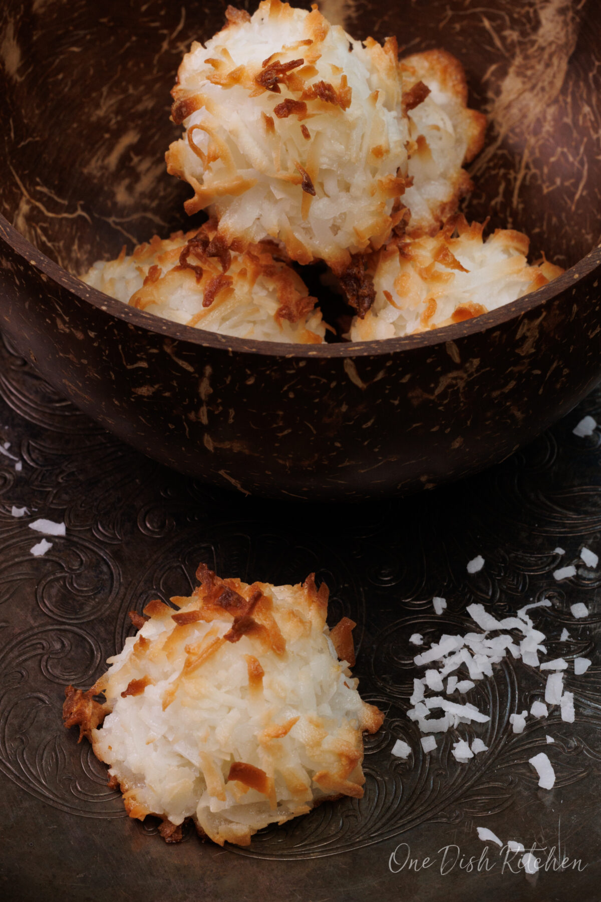coconut macaroons on a silver try next to coconut flakes scattered around the cookie