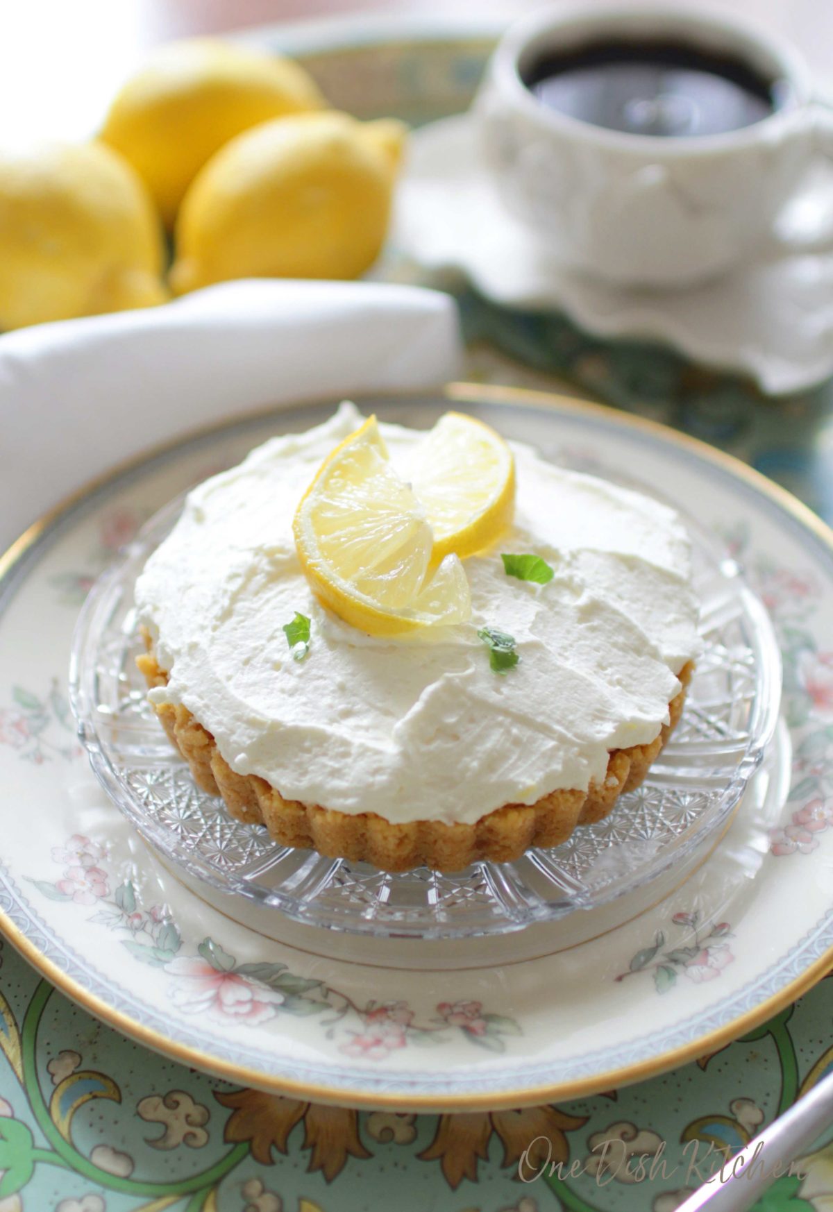 Lemon cheesecake mousse garnished with thin lemon slices on a glass plate with three whole lemons in the background on a metal tray