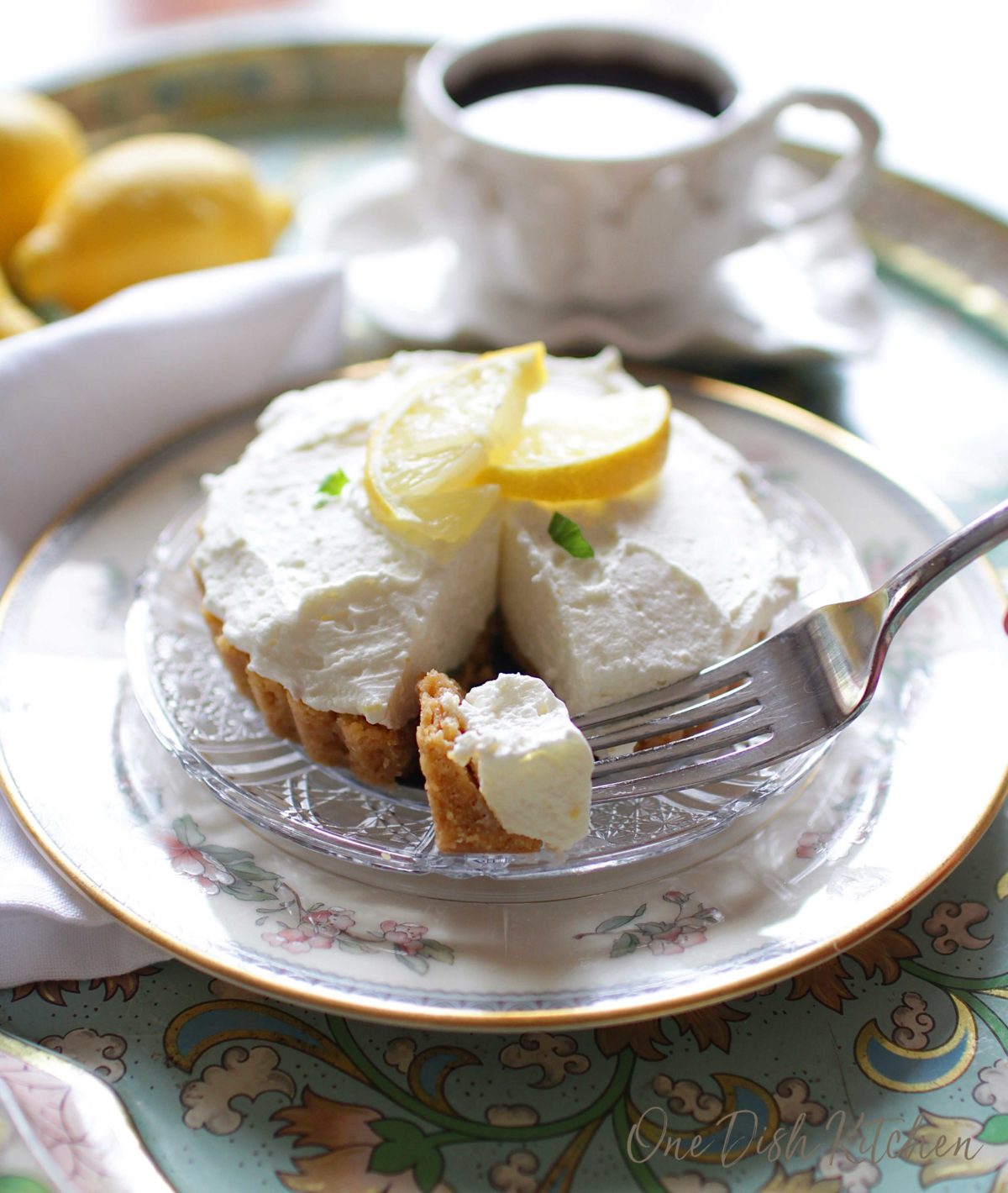 Forkful of lemon cheesecake mousse on a glass plate with whole lemons and a mug of coffee on a metal tray