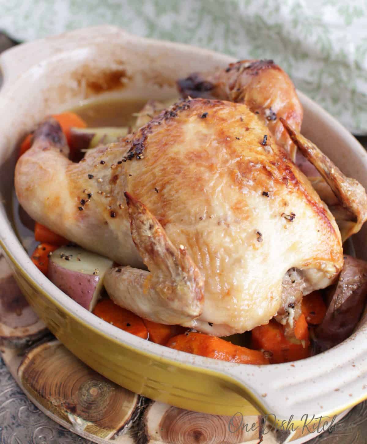 Roasted Cornish Hen on a bed of red potatoes and carrots in a baking dish placed on a wooden trivet that is on a metal tray