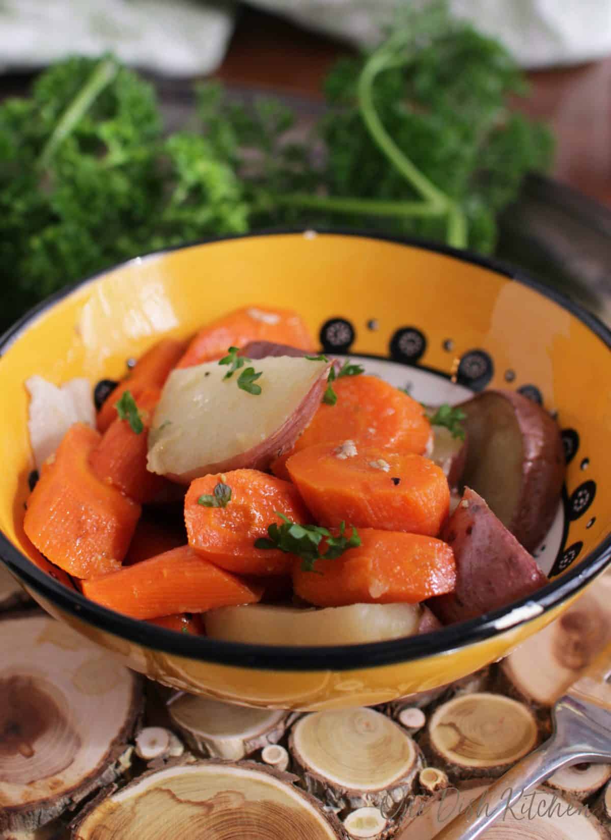 Roasted sliced red potatoes and carrots in a small bowl on a wooden trivet with a fork. 