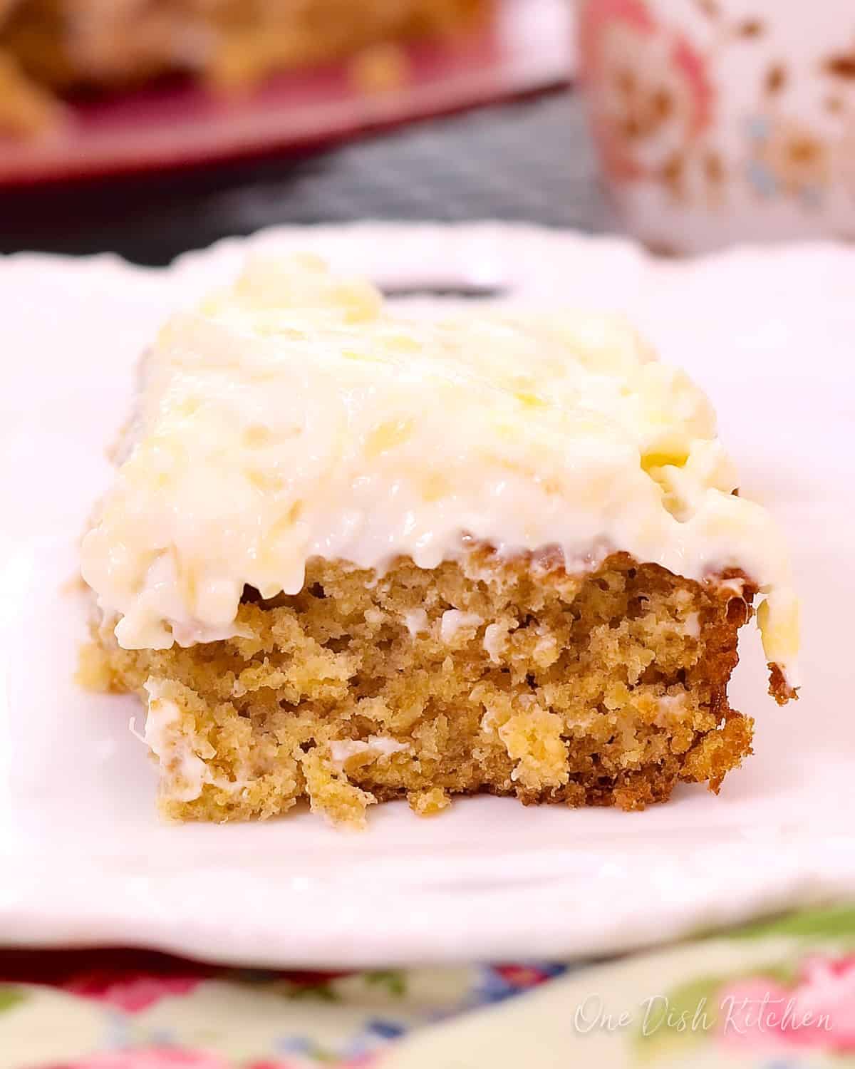 Closeup of a slice of pineapple oat cake with cream cheese frosting on a plate with a floral napkin and a coffee mug in the background.