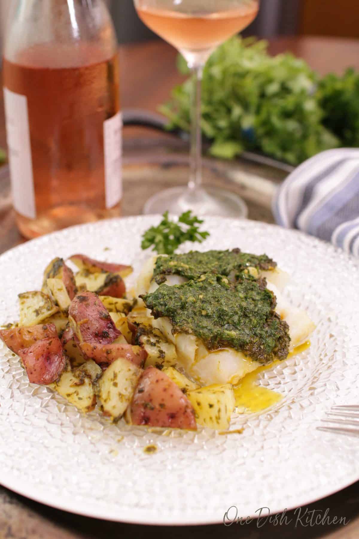 a piece of cod on a plate next to roasted chopped potatoes and a bottle of rose.