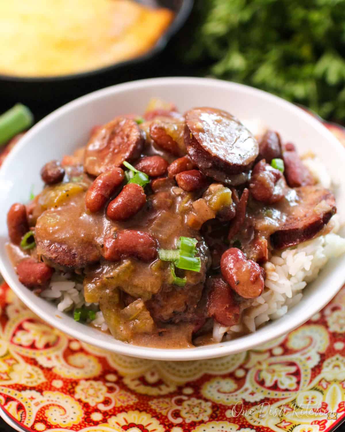 A closeup of a bowl of red beans and white rice with sausage on a tray next to a cast iron skillet of cornbread and a bunch of parsley.