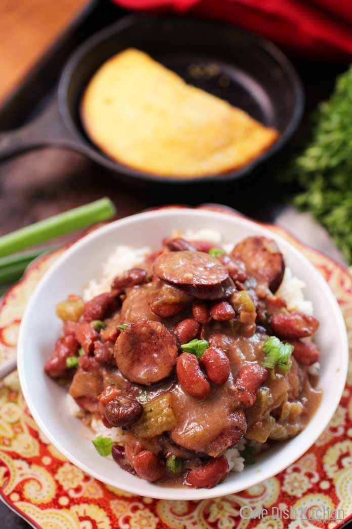 A bowl of sausage and red beans on top of white rice in a bowl next to cornbread in a small skillet.