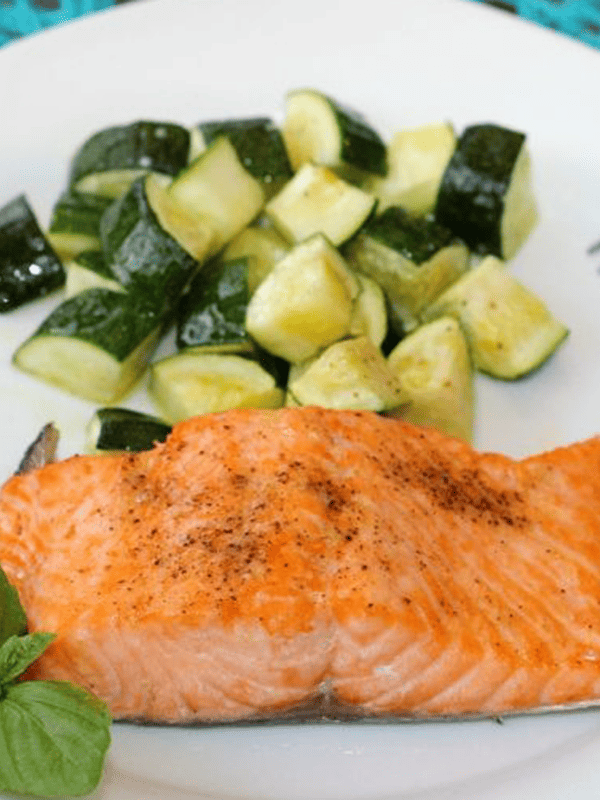 a cooked salmon fillet next to cooked zucchini on a white plate next to fresh basil.