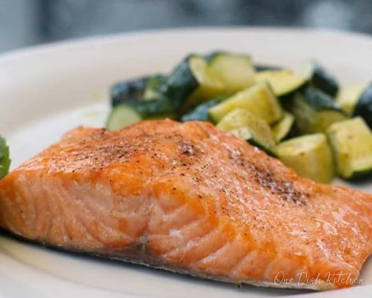 a slice of broiled salmon on a white plate next to cooked zucchini.