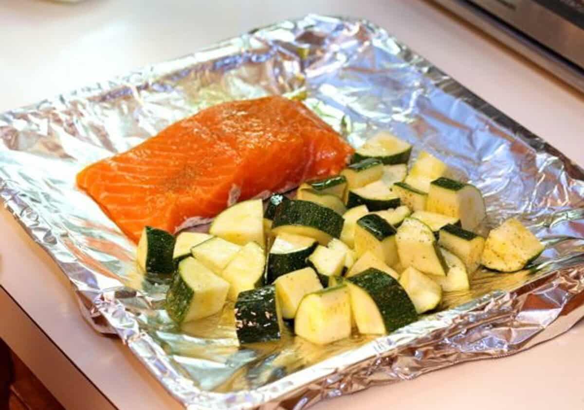 Seasoning raw salmon and zucchini with olive oil, salt, and pepper on a foil-lined toaster oven baking sheet.