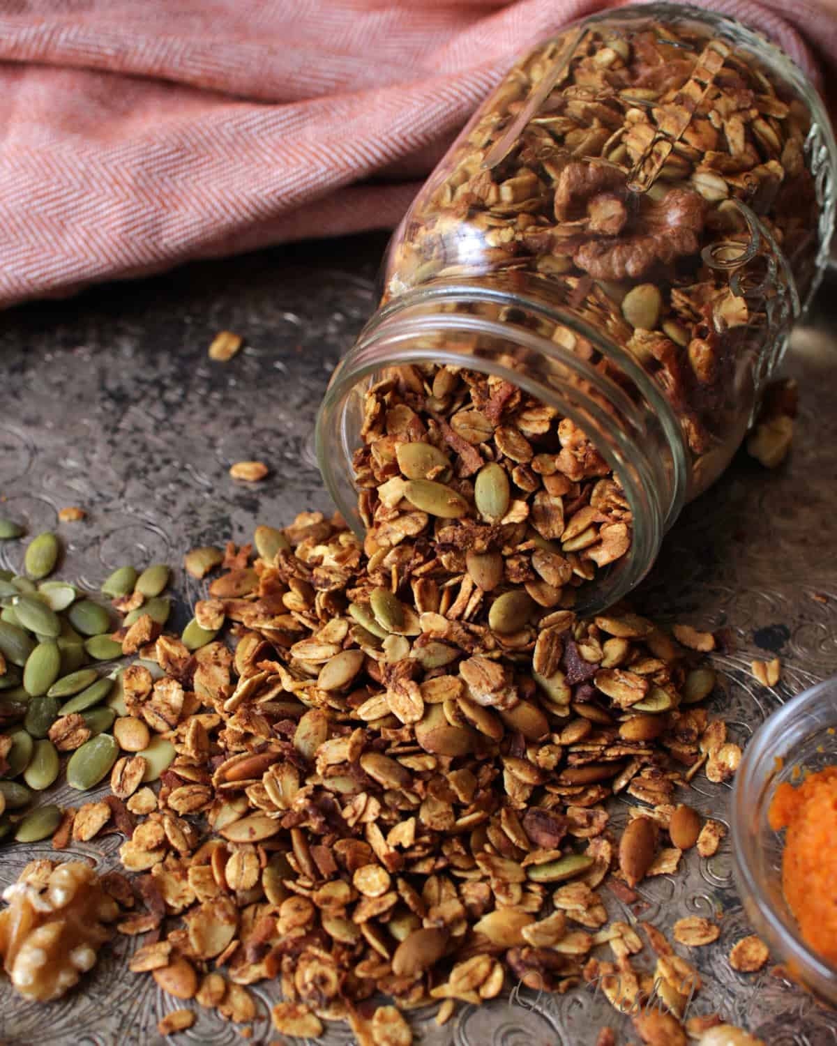a jar of granola spilled out over a silver tray next to raw pumpkin seeds and a bowl of pumpkin puree.