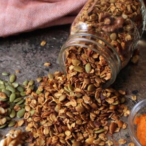a jar of granola spilled out over a silver tray next to raw pumpkin seeds and a bowl of pumpkin puree.
