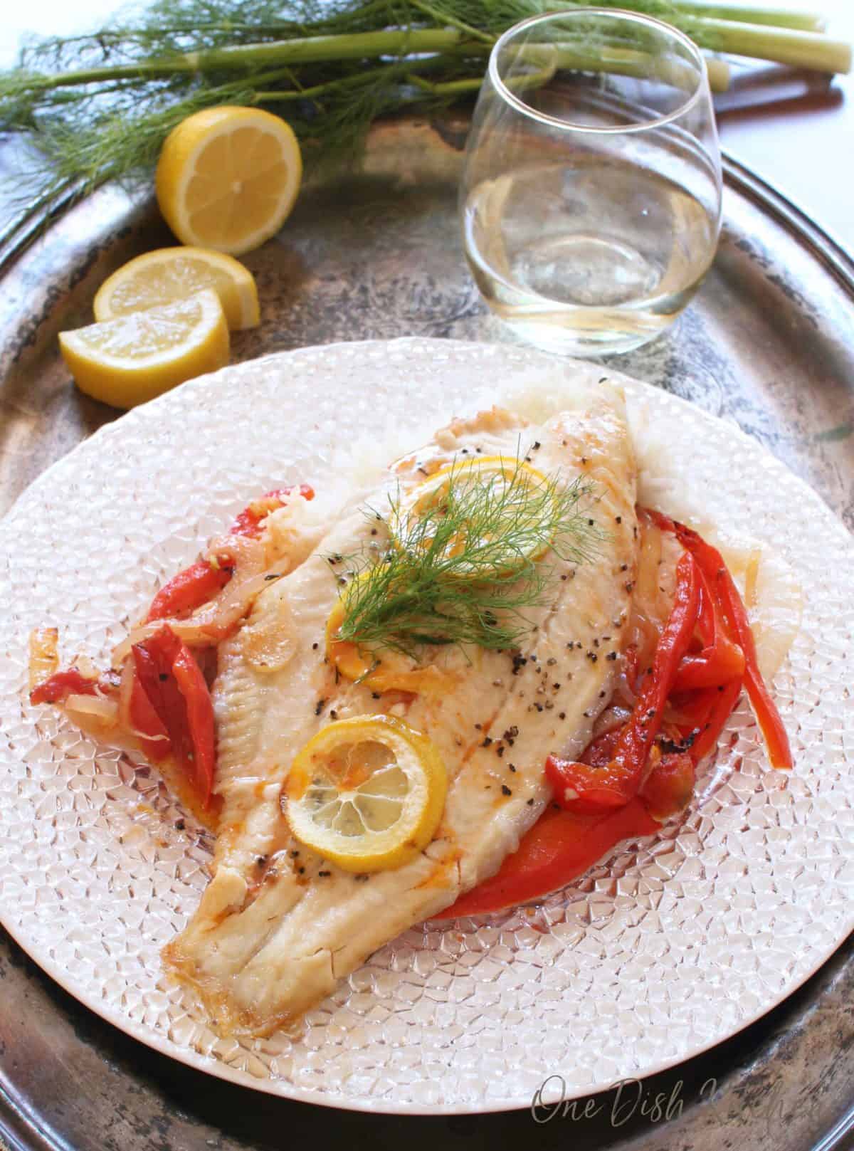 Baked catfish garnished with three lemon wheels over cooked white rice, red peppers, and onions on a glass plate on a metal tray with lemon slices and a glass of white wine.