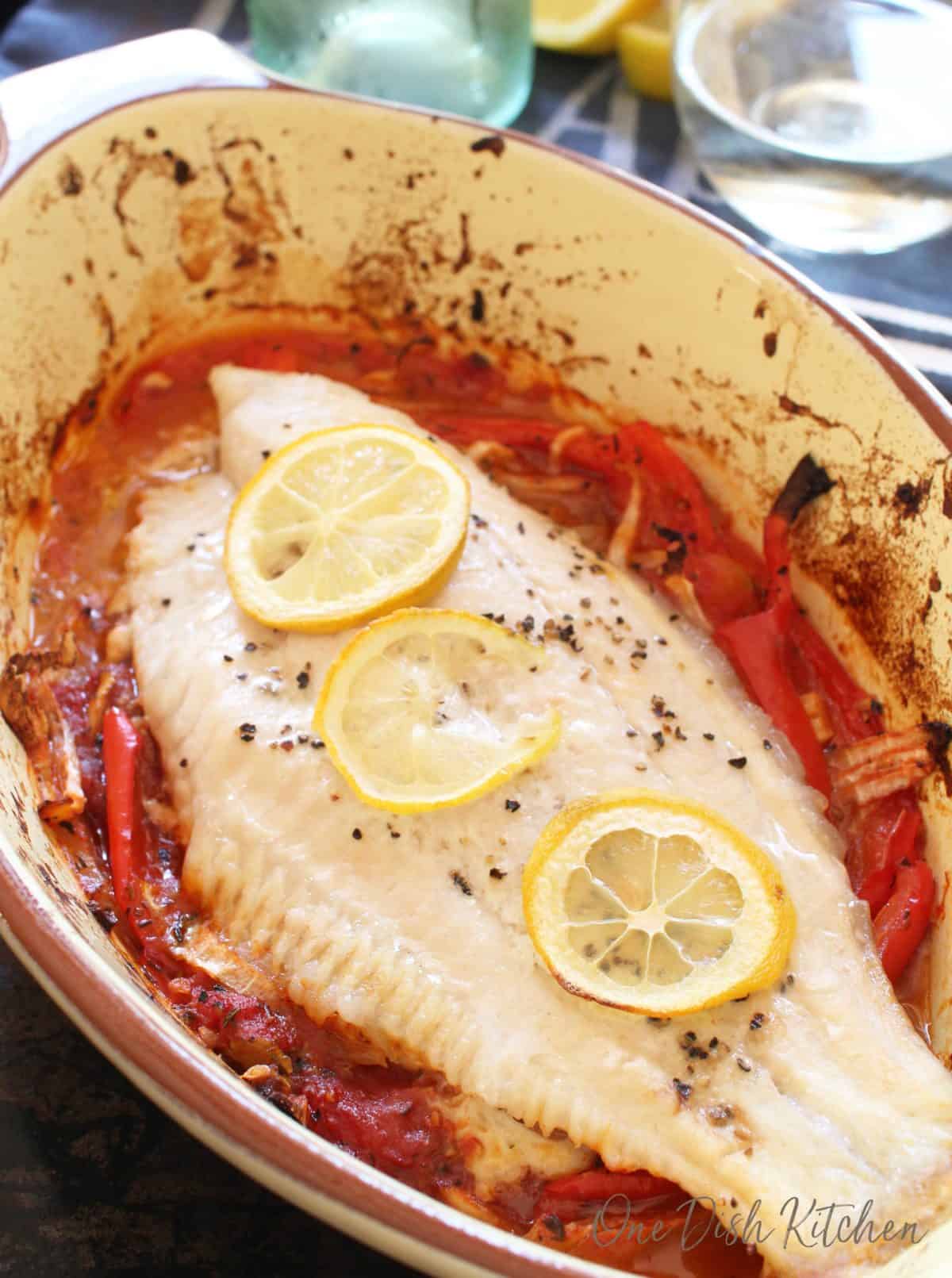 Closeup of catfish in a baking dish garnished with three lemon wheels and pepper flakes baked on a bed of red peppers and onions and there is a glass of white wine in the background.