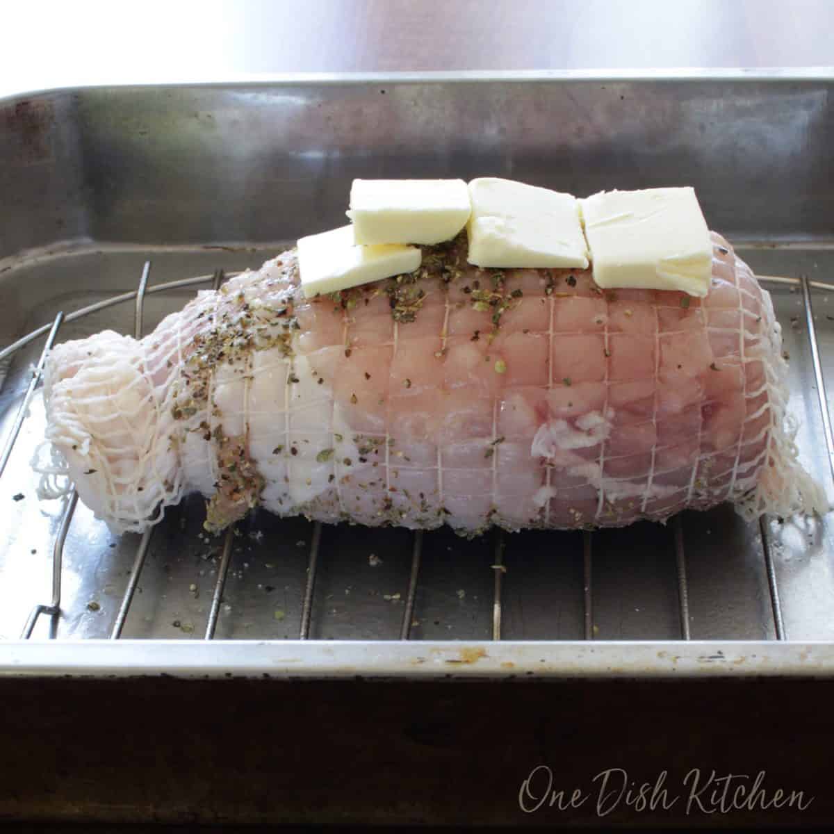 a raw turkey roast in a roasting pan with 4 pieces of butter spread over the top.